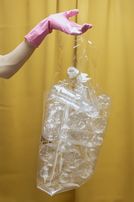 A person wearing a pink rubber glove is holding a large, transparent plastic bag filled with clear plastic bottles and containers, positioned against a yellow curtain background. The bottles inside the bag are empty, varying in size and shape, with some caps visible. The plastic bag appears crumpled and loosely packed, emphasizing the different types of plastic waste collected for disposal or recycling. The scene exemplifies the process of managing waste, highlighting the importance of separate collection for plastics as part of a rubbish clearance service. The lighting is even, with no shadows, allowing for clear visibility of the plastic materials and their textures. This visual represents the step of consolidating recyclables before transportation, supporting the broader context of waste management and waste clearance work that Waste Clearance Stockwell offers for private or alternative disposal methods.