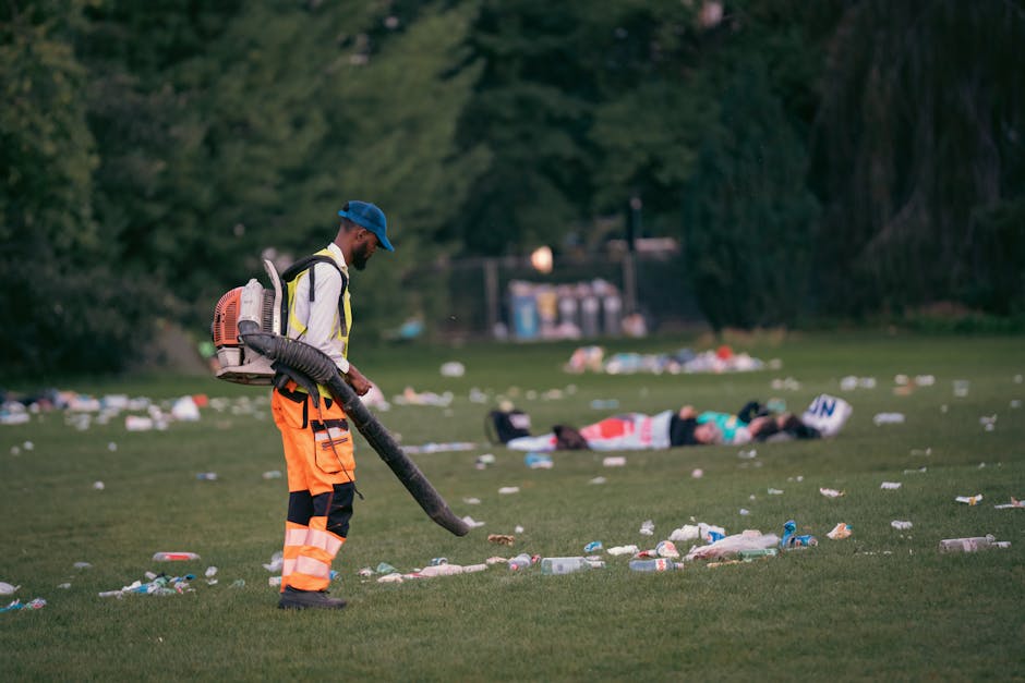 A man dressed in high-visibility orange and black trousers, a white long-sleeved shirt, a blue cap, and carrying a backpack sprayer is using a leaf blower to clear litter from a grassy park area. The ground is scattered with various types of trash, including plastic bottles, paper, and other debris, which are being blown across the scene. In the background, several people are lying on the grass, surrounded by more litter, with tall trees providing a green backdrop. The environment appears to be an outdoor public space, possibly part of a waste clearance or street cleaning effort. The scene highlights the importance of rubbish removal services, such as those offered by Waste Clearance Stockwell, for maintaining cleanliness in communal outdoor areas while demonstrating on-site waste management and disposal activities.