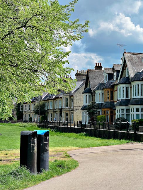 A row of traditional terraced houses with bay windows and pitched roofs featuring chimneys, situated behind a green lawn and a black metal fence. In the foreground, a paved pathway runs parallel to the houses, with two black rubbish bins placed on the grass near the walkway. On the left side, a large deciduous tree with light green leaves extends its branches over the scene, partially shading the pathway. The sky above is partly cloudy with patches of blue visible between the clouds, creating a well-lit outdoor setting typical of residential areas where private waste collection or on-site rubbish disposal may occur, as supported by the presence of street-side bins. The environment appears tidy and well-maintained, reflecting a peaceful suburban neighborhood.