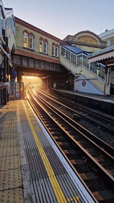 The image depicts an underground railway station platform at Stockwell with train tracks curving towards an illuminated tunnel in the distance. The platform surface is textured with tactile paving in yellow and dark grey, designed for accessibility, and features a yellow safety line along the edge. On the right side, a staircase with a metal handrail leads up to a footbridge passing over the tracks, with the station's brick and concrete structure visible above. The station building has arched windows with brick arch accents and a red brick façade, contributing to the traditional architectural style. A metal bridge spans across the tracks, supporting the station infrastructure, and a small roundel sign with the London Underground logo is mounted near the staircase. The lighting is natural, suggesting the photograph was taken during golden hour, casting a warm glow through the tunnel's opening and creating long shadows. This scene emphasizes the typical infrastructure of an urban underground station, where different elements are arranged to facilitate passenger movement and safety, supporting the context of efficient transportation and potentially related to public or private waste management for station facilities, as managed by Waste Clearance Stockwell.