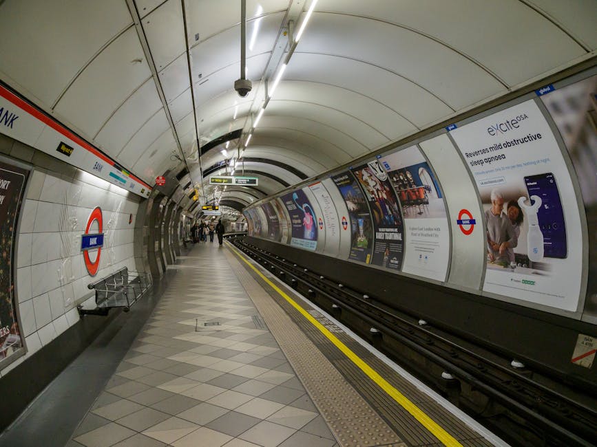 A view of a relatively quiet London Underground station platform adjacent to the Stockwell tube station, with a curved, white-tiled wall on the left side featuring the iconic roundel logo. The platform surface is tiled with light and dark grey geometric patterns, and a yellow line runs along the edge near the tracks for safety. Black train tracks with evenly spaced white fastenings extend into the distance, bordered on the right by a curved, illuminated wall with several large advertising posters depicting various products and services, including a medical device for sleep aids. Several passengers are seen standing and walking along the platform, dressed in casual attire, with some facing the wall while others walk towards the station exit. Overhead, fluorescent lighting fixtures are mounted on the curved ceiling, providing bright, even illumination across the platform. The scene captures a typical scene of an urban underground railway station, indicating efficient public transportation infrastructure, with the setting subtly illustrating the context of local transit rather than private waste or rubbish collection services, aligning with imagery of interior spaces associated with city commuting environments.