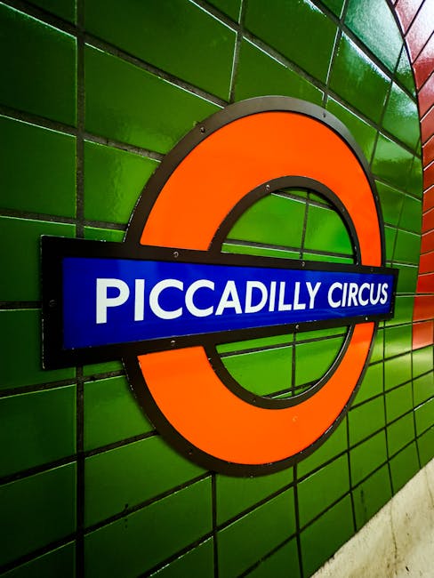 The image depicts an underground railway station platform at Stockwell with train tracks curving towards an illuminated tunnel in the distance. The platform surface is textured with tactile paving in yellow and dark grey, designed for accessibility, and features a yellow safety line along the edge. On the right side, a staircase with a metal handrail leads up to a footbridge passing over the tracks, with the station's brick and concrete structure visible above. The station building has arched windows with brick arch accents and a red brick façade, contributing to the traditional architectural style. A metal bridge spans across the tracks, supporting the station infrastructure, and a small roundel sign with the London Underground logo is mounted near the staircase. The lighting is natural, suggesting the photograph was taken during golden hour, casting a warm glow through the tunnel's opening and creating long shadows. This scene emphasizes the typical infrastructure of an urban underground station, where different elements are arranged to facilitate passenger movement and safety, supporting the context of efficient transportation and potentially related to public or private waste management for station facilities, as managed by Waste Clearance Stockwell.