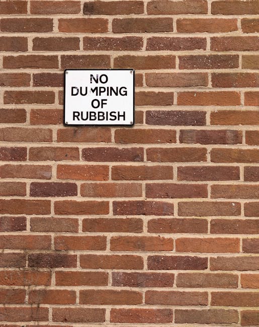 A rectangular white metal sign with black text reading 'NO DUMPING OF RUBBISH' is mounted on a red brick wall with a traditional running bond pattern. The bricks have a mix of reddish-brown and dark brown tones, with light-colored mortar joints in between. The sign is positioned in the upper left section of the image, with the brick wall extending both above and below it, creating a uniform background. The wall appears to be part of an exterior building surface, possibly in an urban or residential area. The lighting is flat, with minimal shadows, highlighting the texture of the brick surface and the sign's clean, reflective surface. This scene, with the clear signage, hints at a designated area for waste management or private disposal, aligning with services provided by Waste Clearance Stockwell in relation to alternative rubbish collection or on-site waste clearance.