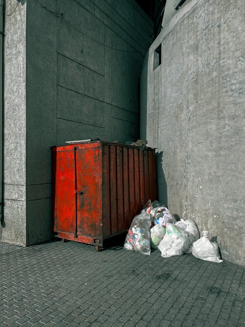 An outdoor urban scene showing a large, weathered red metal waste skip placed against a dark green wall on the left and a plain light grey wall on the right. The skip's surface exhibits patches of rust and chipped paint, indicating exposure to the elements. To the right of the skip, several white plastic rubbish bags filled with mixed household waste are piled on the paved ground, some leaning against the skip. The paving consists of dark, textured concrete slabs arranged in a grid pattern. In the background, the dark green wall extends upward, meeting a small, rectangular window near the top right corner, above the waste. The scene is lit with diffuse natural light, creating soft shadows that enhance the textures of the metal, plastic, and concrete surfaces. This setup suggests a private site or alleyway where Waste Clearance Stockwell provides waste management and alternative rubbish collection services outside standard municipal disposal options, supporting on-site clearance or independent waste handling approaches.